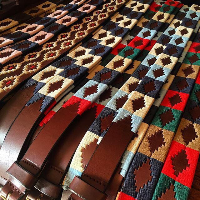 A row of colorful woven leather belts with geometric diamond patterns in various color combinations, including red, blue, beige, brown, green, and black, neatly arranged side by side.”