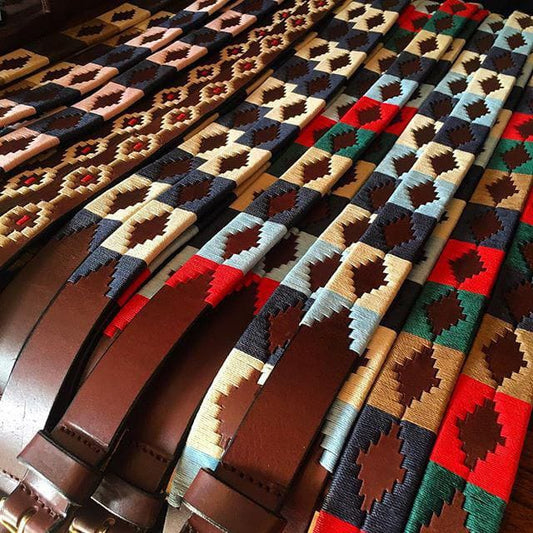 A row of colorful woven leather belts with geometric diamond patterns in various color combinations, including red, blue, beige, brown, green, and black, neatly arranged side by side.”